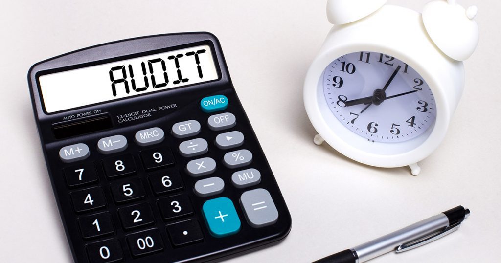 Uma calculadora com a palavra “audit” escrita no visor. Ao lado estão um relógio marcando pouco mais de 8 horas e, mais abaixo, uma caneta de clique.