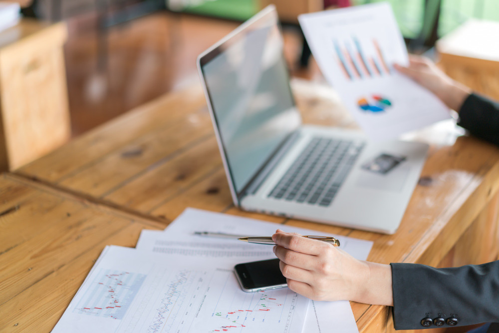 pessoa trabalhando em uma mesa com notebook aberto e segurando papéis com gráficos de balanço.
